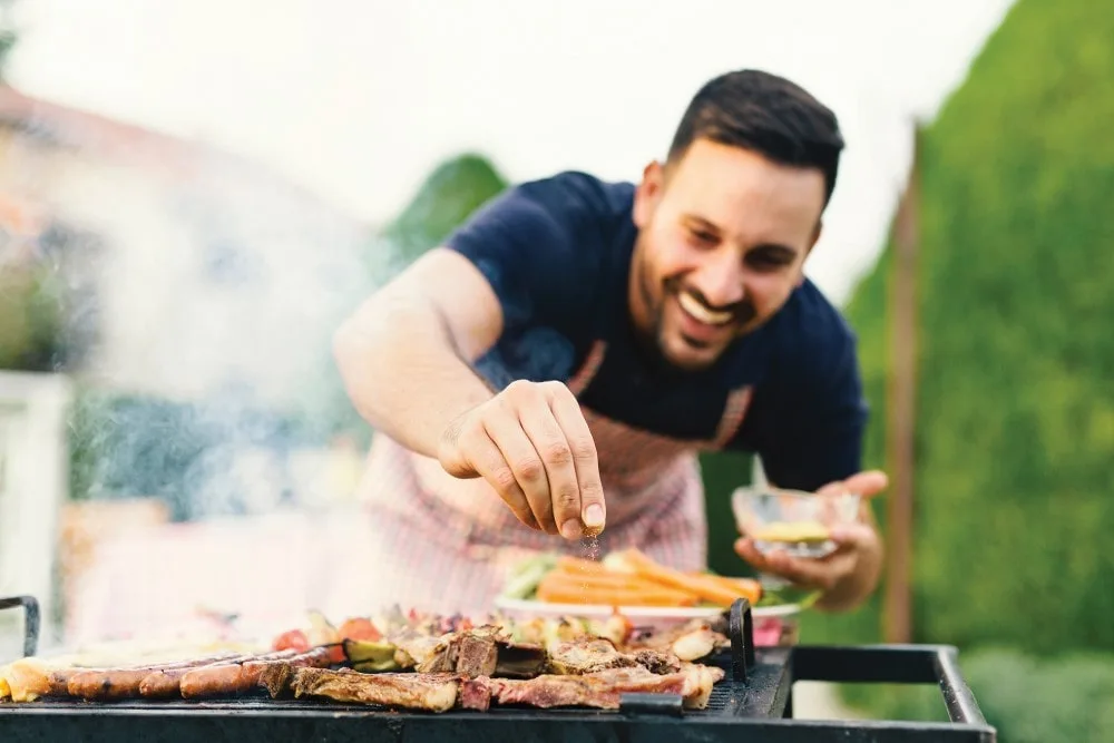 Man seasoning ribs on a grill smiling. He is wearing a plaid red and green apron. He holds his marinade seasoning bowl in his left hand and enthusiastically leans over sprinkling the seasonings with his right.