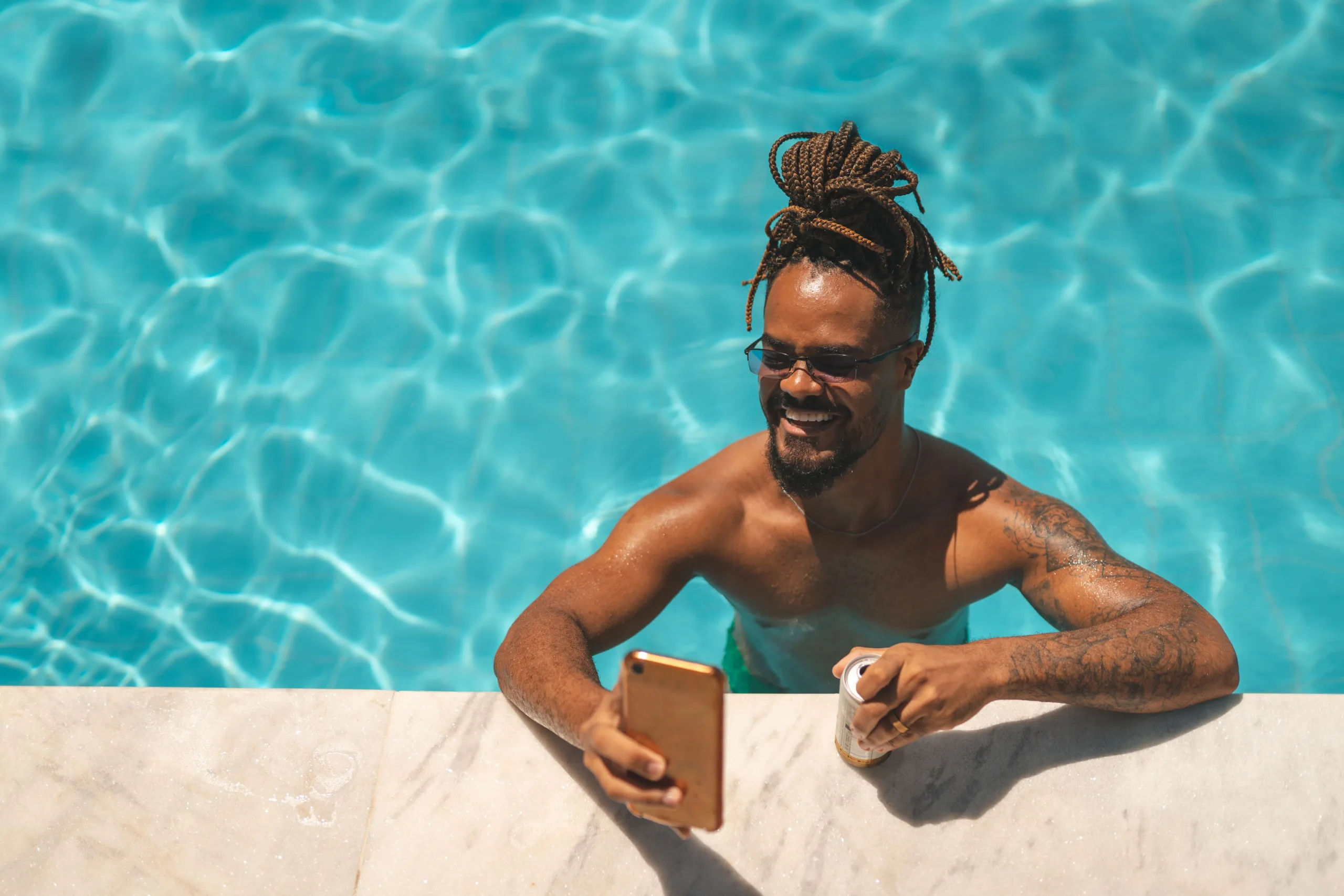 Man with goatee and indistinct tattoos on his left arm smiling in the pool on his phone and resting his arm and drink on the edge of the pool deck.