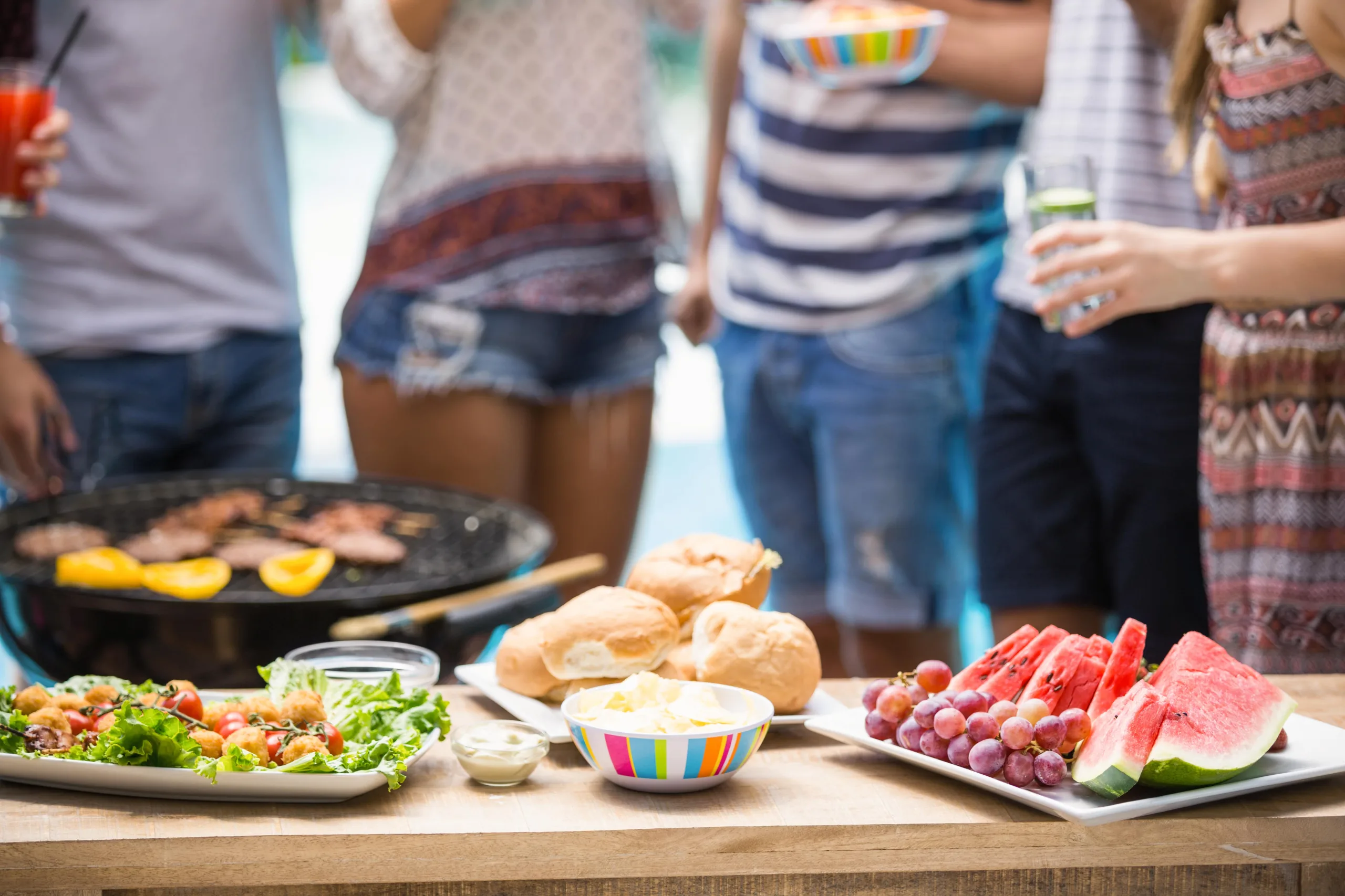 Friends gathered around food on a table with a pool in the background and all holding refreshments. the food is fresh-cut watermelon, grapes, rolls and spread, and a salad and a man is tending to a charcoal grill with bell pepper and burger patties on it.