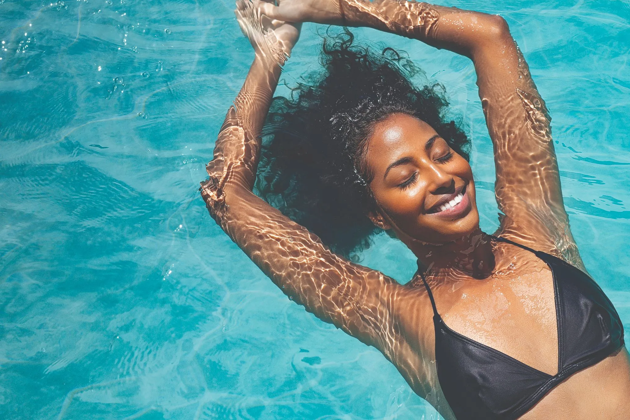 Woman in a black bikini with her arms above her head nestled on top of her curly black hair floating on her back in the pool. Her eyes are closed, and she is smiling.